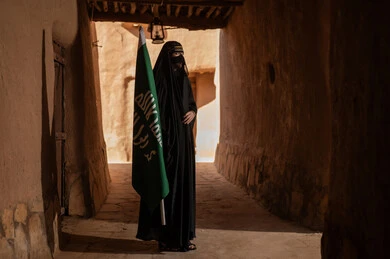 امرأة عربية خليجية سعودية ترتدي العباءة السوداء والبرقع في منزل قديم تمسك بيدها راية التأسيس، ارتداء الزي الشعبي في ذكرى تأسيس الدولة السعودية الاولى 22 فبراير، الفخر والاعتزاز بالهوية الوطنية، نمط حياة البدو الشعبية في البيوت الطينية التراثية، يوم بدينا 1727 م