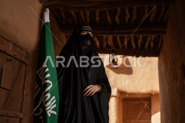 يوم بدينا 1727 م، امرأة عربية خليجية سعودية ترتدي العباءة السوداء والبرقع في منزل قديم تمسك بيدها راية التأسيس، ارتداء الزي الشعبي في ذكرى تأسيس الدولة السعودية الاولى 22 فبراير، الفخر والاعتزاز بالهوية الوطنية، نمط حياة البدو الشعبية في البيوت الطينية التراثية