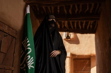 يوم بدينا 1727 م، امرأة عربية خليجية سعودية ترتدي العباءة السوداء والبرقع في منزل قديم تمسك بيدها راية التأسيس، ارتداء الزي الشعبي في ذكرى تأسيس الدولة السعودية الاولى 22 فبراير، الفخر والاعتزاز بالهوية الوطنية، نمط حياة البدو الشعبية في البيوت الطينية التراثية