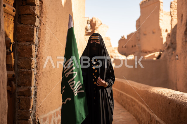 امرأة عربية خليجية سعودية ترتدي العباءة السوداء والبرقع في منزل قديم تمسك بيدها راية التأسيس، ارتداء الزي الشعبي في ذكرى تأسيس الدولة السعودية الاولى 22 فبراير، الفخر والاعتزاز بالهوية الوطنية، نمط حياة البدو الشعبية في البيوت الطينية التراثية، يوم بدينا 1727 م