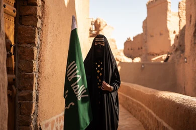 امرأة عربية خليجية سعودية ترتدي العباءة السوداء والبرقع في منزل قديم تمسك بيدها راية التأسيس، ارتداء الزي الشعبي في ذكرى تأسيس الدولة السعودية الاولى 22 فبراير، الفخر والاعتزاز بالهوية الوطنية، نمط حياة البدو الشعبية في البيوت الطينية التراثية، يوم بدينا 1727 م