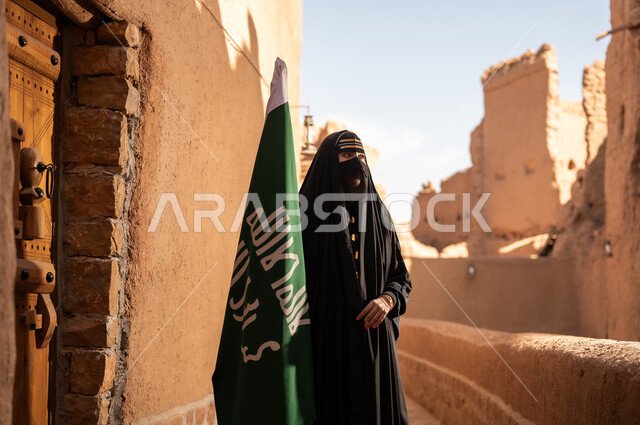 امرأة عربية خليجية سعودية ترتدي العباءة السوداء والبرقع في منزل قديم تمسك بيدها راية التأسيس، ارتداء الزي الشعبي في ذكرى تأسيس الدولة السعودية الاولى 22 فبراير، الفخر والاعتزاز بالهوية الوطنية، نمط حياة البدو الشعبية في البيوت الطينية التراثية، يوم بدينا 1727 م