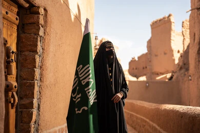 امرأة عربية خليجية سعودية ترتدي العباءة السوداء والبرقع في منزل قديم تمسك بيدها راية التأسيس، ارتداء الزي الشعبي في ذكرى تأسيس الدولة السعودية الاولى 22 فبراير، الفخر والاعتزاز بالهوية الوطنية، نمط حياة البدو الشعبية في البيوت الطينية التراثية، يوم بدينا 1727 م