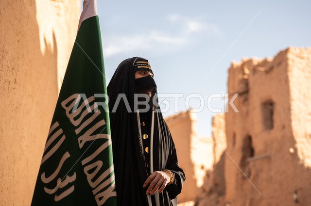 يوم بدينا 1727 م، امرأة عربية خليجية سعودية ترتدي العباءة السوداء والبرقع في منزل قديم تمسك بيدها راية التأسيس، ارتداء الزي الشعبي في ذكرى تأسيس الدولة السعودية الاولى 22 فبراير، الفخر والاعتزاز بالهوية الوطنية، نمط حياة البدو الشعبية في البيوت الطينية التراثية