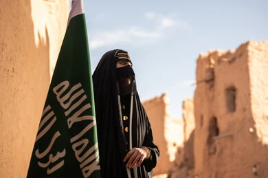 يوم بدينا 1727 م، امرأة عربية خليجية سعودية ترتدي العباءة السوداء والبرقع في منزل قديم تمسك بيدها راية التأسيس، ارتداء الزي الشعبي في ذكرى تأسيس الدولة السعودية الاولى 22 فبراير، الفخر والاعتزاز بالهوية الوطنية، نمط حياة البدو الشعبية في البيوت الطينية التراثية