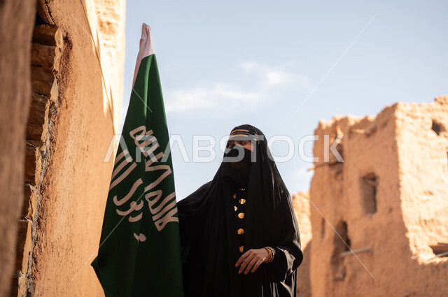 On the day we began, 1727 AD, a Saudi Arabian Gulf woman wearing a black abaya and burqa in an old house, holding the flag of the founding, wearing traditional dress on the anniversary of the founding of the first Saudi state, February 22, pride and honor in national identity, the popular Bedouin lifestyle in traditional mud houses.