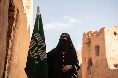 يوم بدينا 1727 م، امرأة عربية خليجية سعودية ترتدي العباءة السوداء والبرقع في منزل قديم تمسك بيدها راية التأسيس، ارتداء الزي الشعبي في ذكرى تأسيس الدولة السعودية الاولى 22 فبراير، الفخر والاعتزاز بالهوية الوطنية، نمط حياة البدو الشعبية في البيوت الطينية التراثية