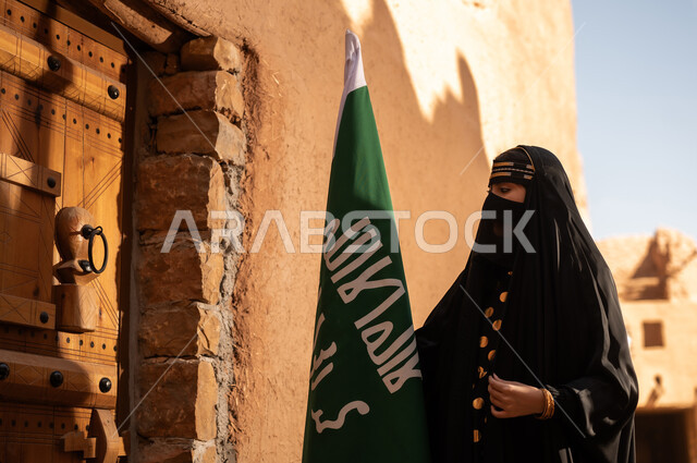 يوم بدينا 1727 م، امرأة عربية خليجية سعودية ترتدي العباءة السوداء والبرقع في منزل قديم تمسك بيدها راية التأسيس، ارتداء الزي الشعبي في ذكرى تأسيس الدولة السعودية الاولى 22 فبراير، الفخر والاعتزاز بالهوية الوطنية، نمط حياة البدو الشعبية في البيوت الطينية التراثية