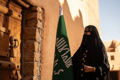 يوم بدينا 1727 م، امرأة عربية خليجية سعودية ترتدي العباءة السوداء والبرقع في منزل قديم تمسك بيدها راية التأسيس، ارتداء الزي الشعبي في ذكرى تأسيس الدولة السعودية الاولى 22 فبراير، الفخر والاعتزاز بالهوية الوطنية، نمط حياة البدو الشعبية في البيوت الطينية التراثية