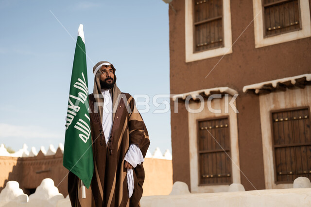 أزياء شعبية سعودية، رجل عربي خليجي سعودي يرتدي زي تراثي سعودي للمناسبات الوطنية يمسك بيده علم التأسيس السعودي، تأسيس المملكة 1727م ،تصميم معماري عربي قديم، بيوت طينية تراثية قديمة ، يوم بدينا ٢٢فبراير
