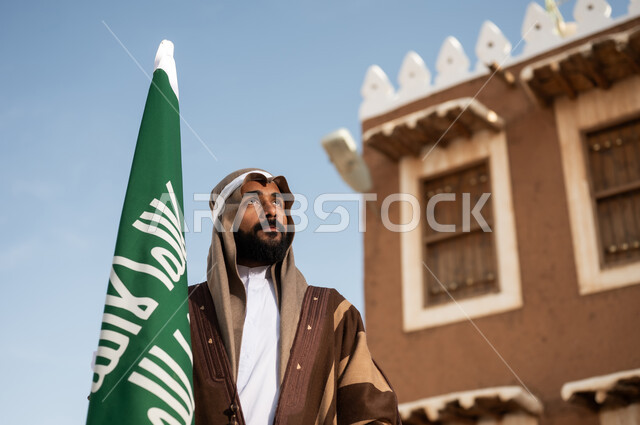 أزياء شعبية سعودية، رجل عربي خليجي سعودي يرتدي زي تراثي سعودي للمناسبات الوطنية يمسك بيده علم التأسيس السعودي، تأسيس المملكة 1727م ،تصميم معماري عربي قديم، بيوت طينية تراثية قديمة ، يوم بدينا ٢٢فبراير