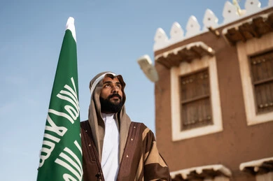 Saudi folk costumes, a Gulf Arab Saudi man wearing a traditional Saudi outfit for national occasions holding the Saudi foundation flag in his hand, the establishment of the kingdom in 1727 AD, ancient Arab architectural design, old traditional mud houses, the day we started on February 22.