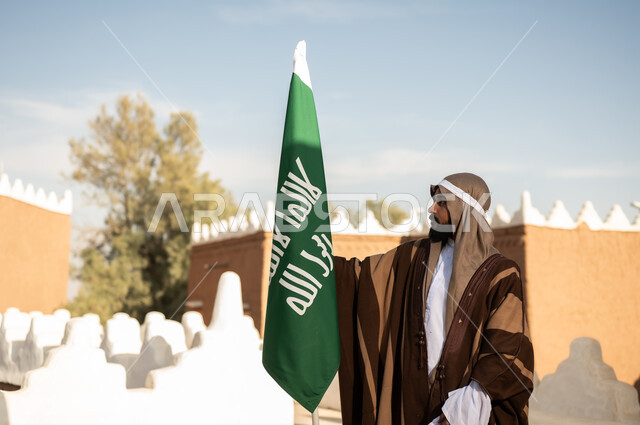 Saudi folk costumes, a Gulf Arab Saudi man wearing a traditional Saudi outfit for national occasions holding the Saudi foundation flag in his hand, the establishment of the kingdom in 1727 AD, ancient Arab architectural design, old traditional mud houses, the day we started on February 22.
