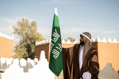 أزياء شعبية سعودية، رجل عربي خليجي سعودي يرتدي زي تراثي سعودي للمناسبات الوطنية يمسك بيده علم التأسيس السعودي، تأسيس المملكة 1727م ،تصميم معماري عربي قديم، بيوت طينية تراثية قديمة ، يوم بدينا ٢٢فبراير