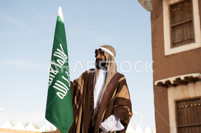 أزياء شعبية سعودية، رجل عربي خليجي سعودي يرتدي زي تراثي سعودي للمناسبات الوطنية يمسك بيده علم التأسيس السعودي، تأسيس المملكة 1727م ،تصميم معماري عربي قديم، بيوت طينية تراثية قديمة ، يوم بدينا ٢٢فبراير
