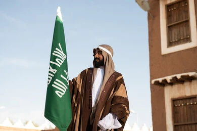 أزياء شعبية سعودية، رجل عربي خليجي سعودي يرتدي زي تراثي سعودي للمناسبات الوطنية يمسك بيده علم التأسيس السعودي، تأسيس المملكة 1727م ،تصميم معماري عربي قديم، بيوت طينية تراثية قديمة ، يوم بدينا ٢٢فبراير