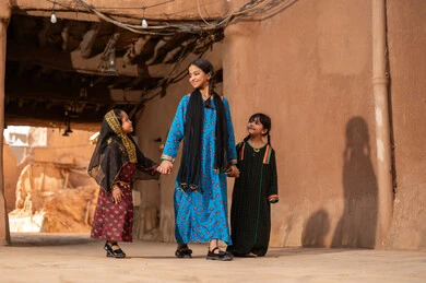 تعابير السرور والسعادة، فتاة عربية خليجية سعودية ترتدي الزي التراثي تمشي مع اختيها في حي طيني قديم, ذكرى تأسيس الدوله السعوديه الاولى على يد الامام محمد بن سعود, يوم بدينا 22 فبراير 1727م, العمق التاريخي والحضاري للمملكة، ارتداء الأزياء التقليدية تعكس التراث