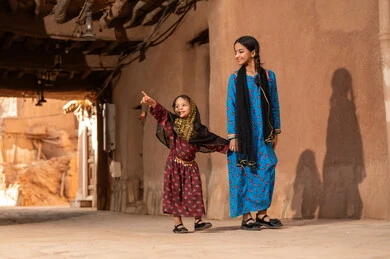 Referring to something, a Gulf Arab Saudi girl wearing traditional attire walking with her sister in an old mud neighborhood, commemorating the founding of the first Saudi state by Imam Muhammad bin Saud, on the day we began, February 22, 1727 AD, the historical and cultural depth of the Kingdom, wearing traditional costumes reflects heritage.
