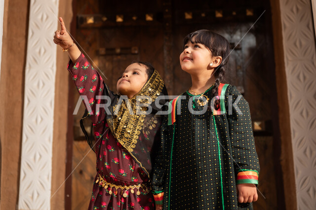 Referring to something, a Gulf Arab Saudi girl wearing traditional attire walking with her sister in an old mud neighborhood, commemorating the founding of the first Saudi state by Imam Muhammad bin Saud, on the day we began, February 22, 1727 AD, the historical and cultural depth of the Kingdom, wearing traditional costumes reflects heritage.