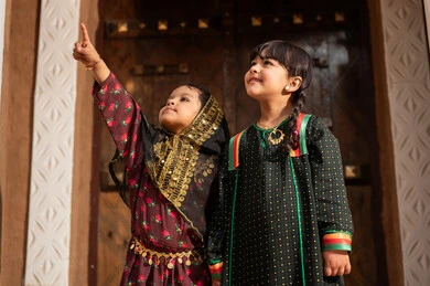 Referring to something, a Gulf Arab Saudi girl wearing traditional attire walking with her sister in an old mud neighborhood, commemorating the founding of the first Saudi state by Imam Muhammad bin Saud, on the day we began, February 22, 1727 AD, the historical and cultural depth of the Kingdom, wearing traditional costumes reflects heritage.