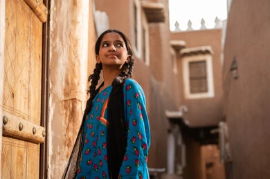 A Gulf Arab Saudi girl wearing traditional attire stands in front of an old wooden door, commemorating the founding of the first Saudi state by Imam Muhammad bin Saud, on the day we began, February 22, 1727. The historical and cultural depth of the Kingdom, wearing traditional costumes reflects the heritage.