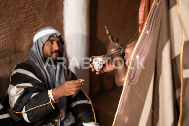 حسن الضيافة والكرم عند البدو، يوم بدينا 1727 م، حي شعبي وبيوت طينية تراثية، ذكرى تأسيس الدولة السعودية الاولى 22 فبراير, إظهار الحب والولاء للدولة والوطن, رجل عربي خليجي سعودي يرتدي زي شعبي تراثي يقوم بتقديم القهوة العربية، فخر واعتزاز بالهوية الوطنية