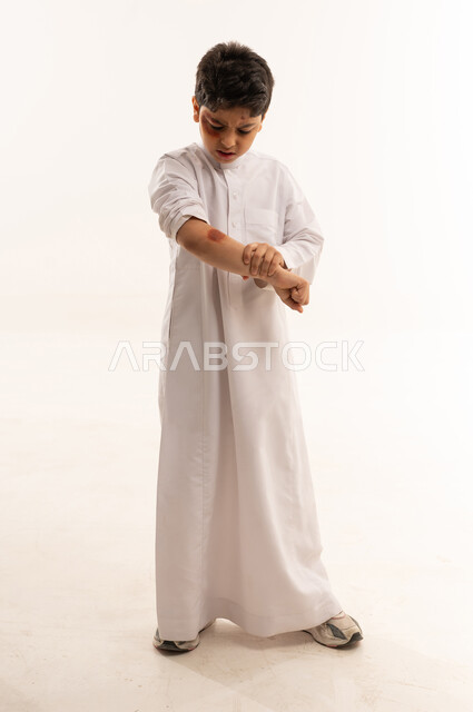 Saudi Boy in Thobe Inspecting Arm Bruise on White Background