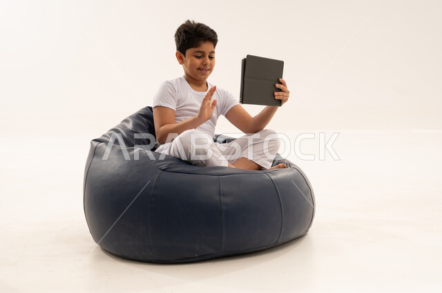 طفل عربي يلوح لجهاز لوحي على مقعد محشو أزرق