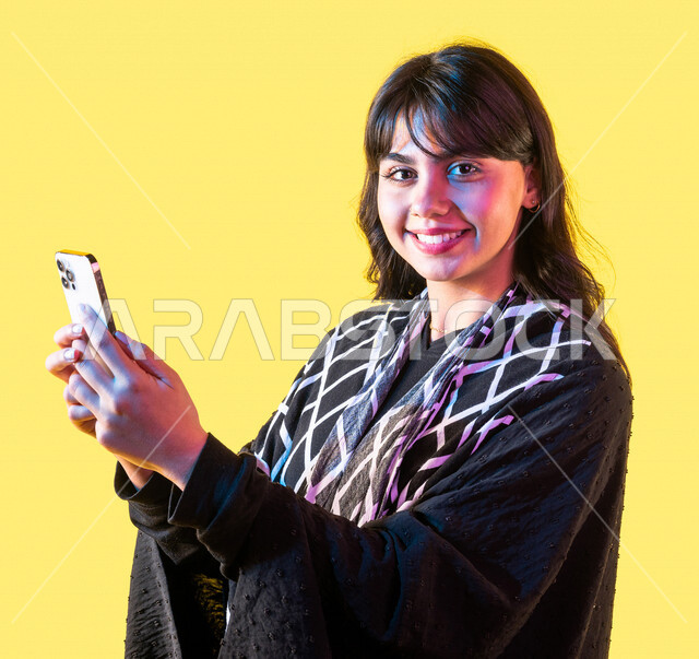 Portrait of a smiling Saudi Gulf young woman, looking at the camera, using a mobile phone, surfing the Internet, e-marketing, using modern technology devices, using social media, communicating with friends, yellow background