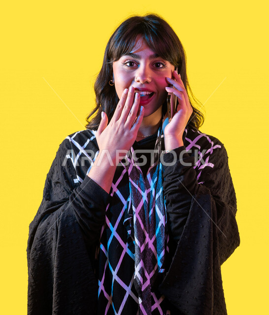 بورتريه لشابة عربية خليجية سعودية ، تقوم بإجراء مكالمة هاتفية عبر الهاتف المحمول ، بإيماءات وجه ويدين مختلفة تدل على الدهشة والسعادة لحصولها على شيء ما ، مفهوم تلقي الأخبار السعيدة ، مفهوم الذهول والإنبهار ، خلفية صفراء