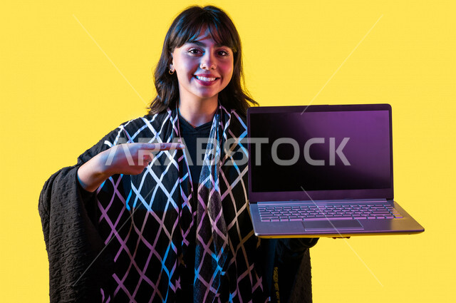 Portrait of a Saudi Gulf woman, using a laptop, displaying a laptop screen, using modern technology devices, a working session, browsing the Internet, displaying services and applications via laptop, digital e-marketing, advertising concept, yellow backgr