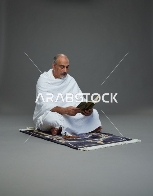 بورتريه لحاج كبير السن عربي سعودي خليجي بملابس الإحرام يقرأ القرآن وبيده مسبحة يجلس على سجادة صلاه, اداء مناسك الحج والعمره , خلفيه رماديه