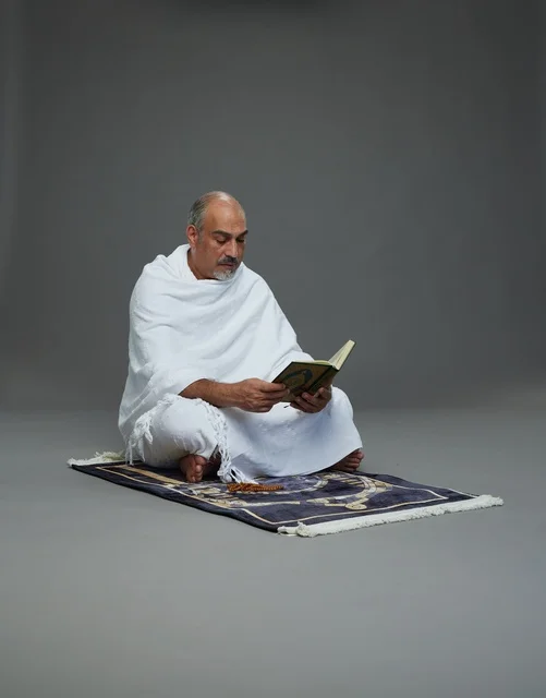 A portrait of an elderly Arab Saudi Gulf pilgrim in Ihram clothing reading the Quran, holding a prayer bead, sitting on a prayer rug, performing the rituals of Hajj and Umrah, with a gray background.