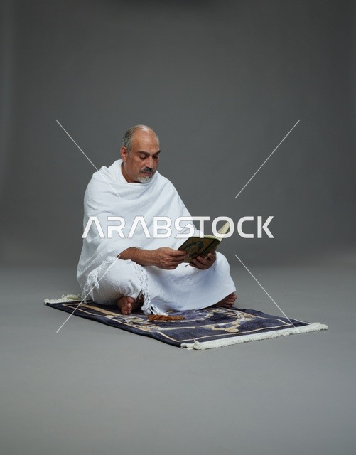 بورتريه لحاج كبير السن عربي سعودي خليجي بملابس الإحرام يقرأ القرآن وبيده مسبحة يجلس على سجادة صلاه, اداء مناسك الحج والعمره , خلفيه رماديه