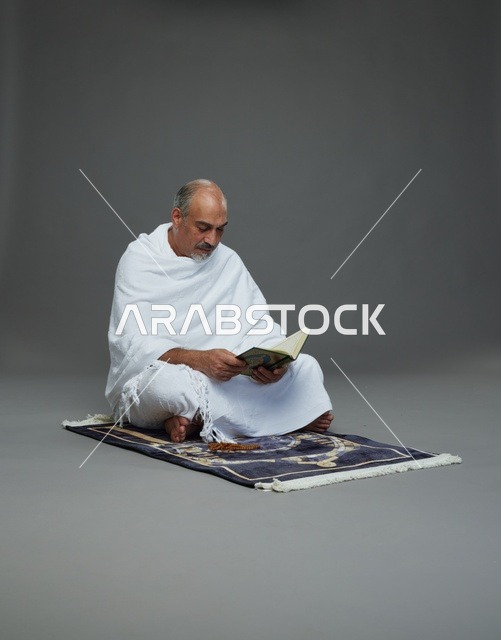 بورتريه لحاج كبير السن عربي سعودي خليجي بملابس الإحرام يقرأ القرآن وبيده مسبحة يجلس على سجادة صلاه, اداء مناسك الحج والعمره , خلفيه رماديه