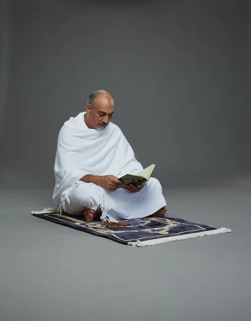 A portrait of an elderly Arab Saudi Gulf pilgrim in Ihram clothing reading the Quran, holding a prayer bead, sitting on a prayer rug, performing the rituals of Hajj and Umrah, with a gray background.