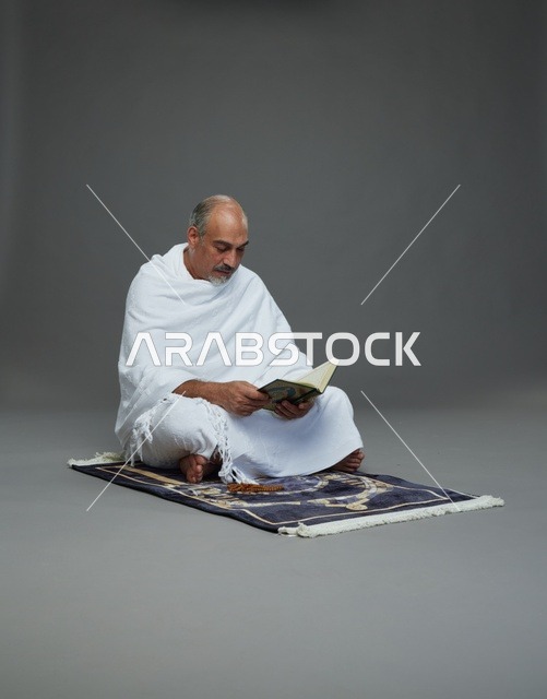 بورتريه لحاج كبير السن عربي سعودي خليجي بملابس الإحرام يقرأ القرآن وبيده مسبحة يجلس على سجادة صلاه, اداء مناسك الحج والعمره , خلفيه رماديه