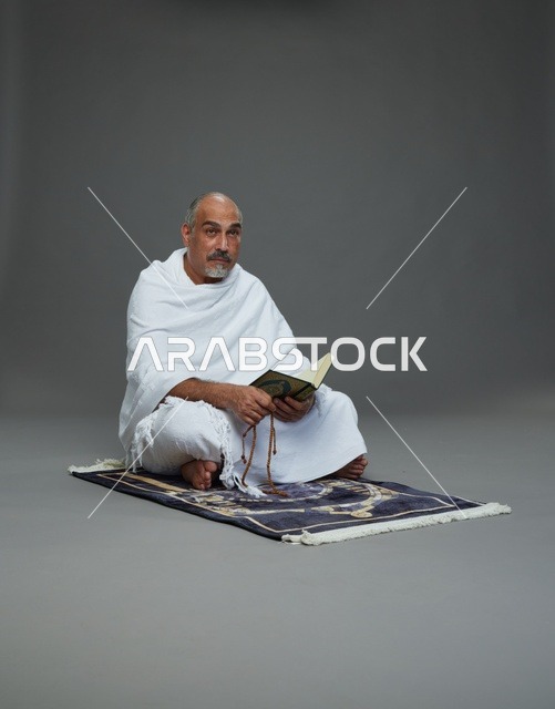 بورتريه لحاج كبير السن عربي سعودي خليجي بملابس الإحرام يقرأ القرآن وبيده مسبحة يجلس على سجادة صلاه, اداء مناسك الحج والعمره , خلفيه رماديه