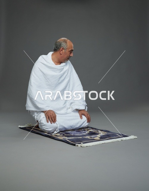 بورتريه لحاج كبير السن عربي سعودي خليجي بملابس الإحرام  يجلس على سجادة صلاه ويصلي, اداء مناسك الحج والعمره , خلفيه رماديه