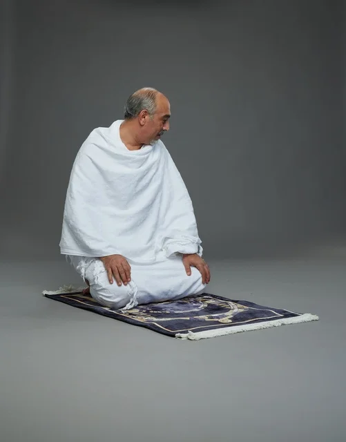 بورتريه لحاج كبير السن عربي سعودي خليجي بملابس الإحرام  يجلس على سجادة صلاه ويصلي, اداء مناسك الحج والعمره , خلفيه رماديه