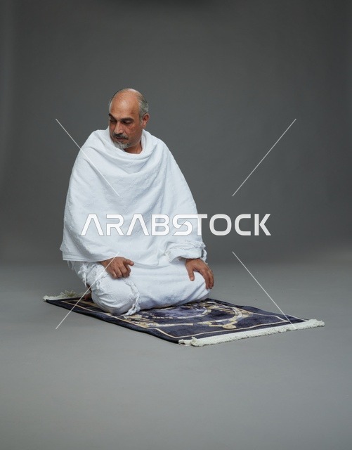 بورتريه لحاج كبير السن عربي سعودي خليجي بملابس الإحرام  يجلس على سجادة صلاه ويصلي, اداء مناسك الحج والعمره , خلفيه رماديه
