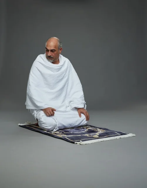 بورتريه لحاج كبير السن عربي سعودي خليجي بملابس الإحرام  يجلس على سجادة صلاه ويصلي, اداء مناسك الحج والعمره , خلفيه رماديه