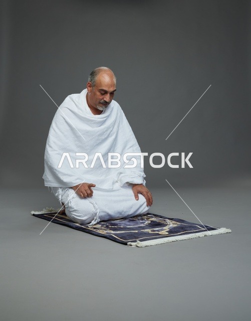 بورتريه لحاج كبير السن عربي سعودي خليجي بملابس الإحرام  يجلس على سجادة صلاه ويصلي, اداء مناسك الحج والعمره , خلفيه رماديه
