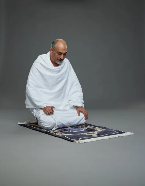 بورتريه لحاج كبير السن عربي سعودي خليجي بملابس الإحرام  يجلس على سجادة صلاه ويصلي, اداء مناسك الحج والعمره , خلفيه رماديه