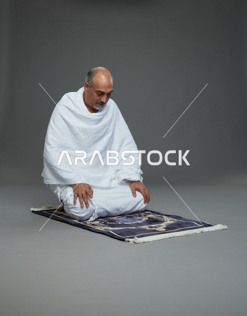 بورتريه لحاج كبير السن عربي سعودي خليجي بملابس الإحرام  يجلس على سجادة صلاه ويصلي, اداء مناسك الحج والعمره , خلفيه رماديه