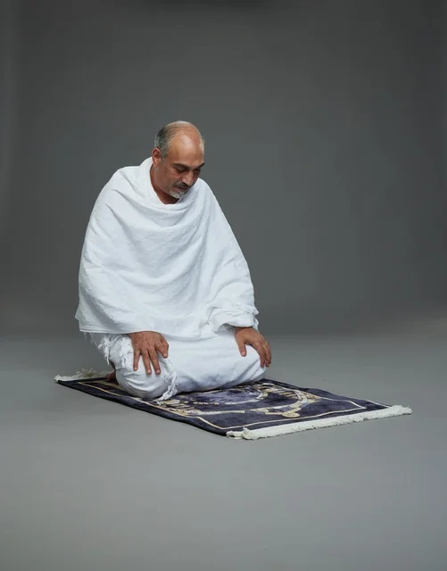 بورتريه لحاج كبير السن عربي سعودي خليجي بملابس الإحرام  يجلس على سجادة صلاه ويصلي, اداء مناسك الحج والعمره , خلفيه رماديه