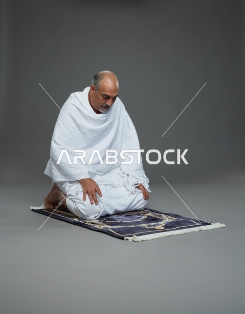 بورتريه لحاج كبير السن عربي سعودي خليجي بملابس الإحرام  يجلس على سجادة صلاه ويصلي, اداء مناسك الحج والعمره , خلفيه رماديه