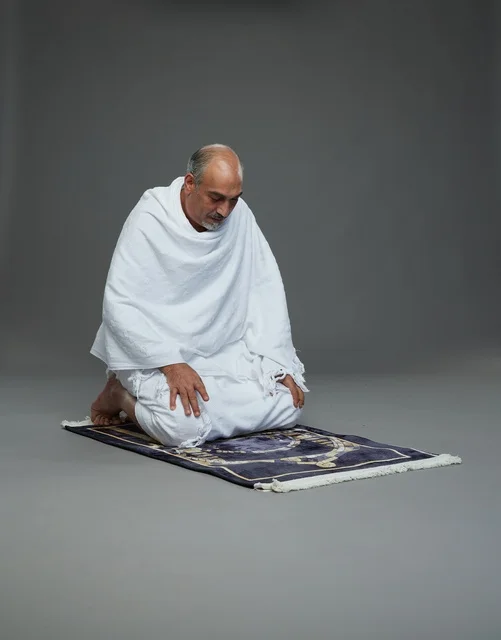 A portrait of an elderly Arab Saudi Gulf pilgrim in Ihram clothing sitting on a prayer rug and praying, performing the rituals of Hajj and Umrah, with a gray background.