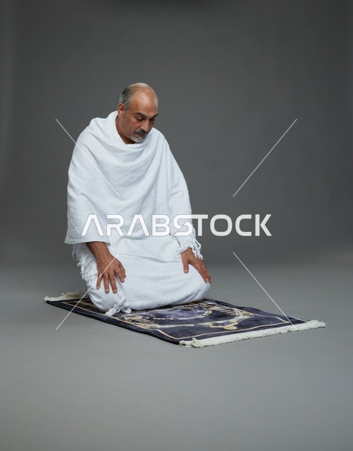 بورتريه لحاج كبير السن عربي سعودي خليجي بملابس الإحرام  يجلس على سجادة صلاه ويصلي, اداء مناسك الحج والعمره , خلفيه رماديه