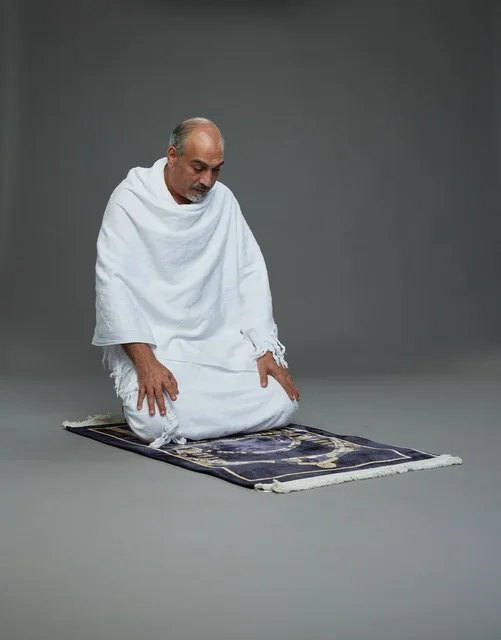 بورتريه لحاج كبير السن عربي سعودي خليجي بملابس الإحرام  يجلس على سجادة صلاه ويصلي, اداء مناسك الحج والعمره , خلفيه رماديه
